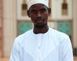 A man in traditional white attire standing in front of architectural details in Abuja, Nigeria.